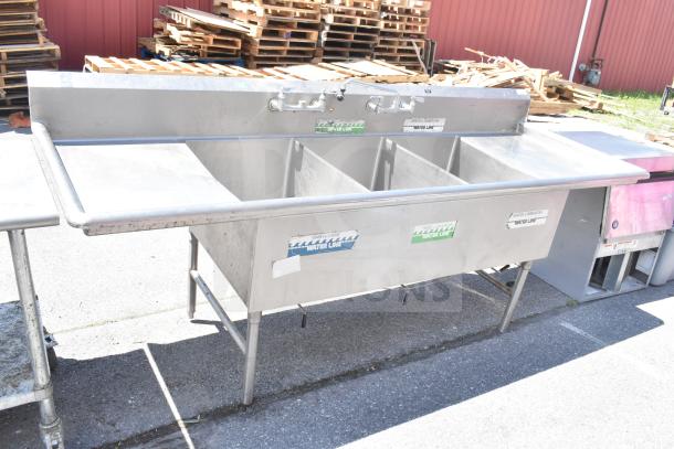 Commercial 3 bay stainless steel sink with dual drain boards, faucet handles, and visible water line labels, good condition.