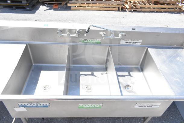 Commercial 3 bay sink with dual drain boards, stainless steel, labels for wash, rinse, sanitize, and visible faucet controls.