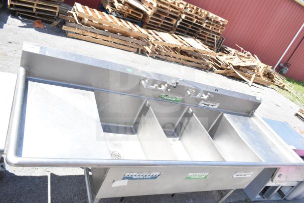 Stainless steel commercial 3 bay sink with dual drain boards, faucets, and visible labels indicating water lines.