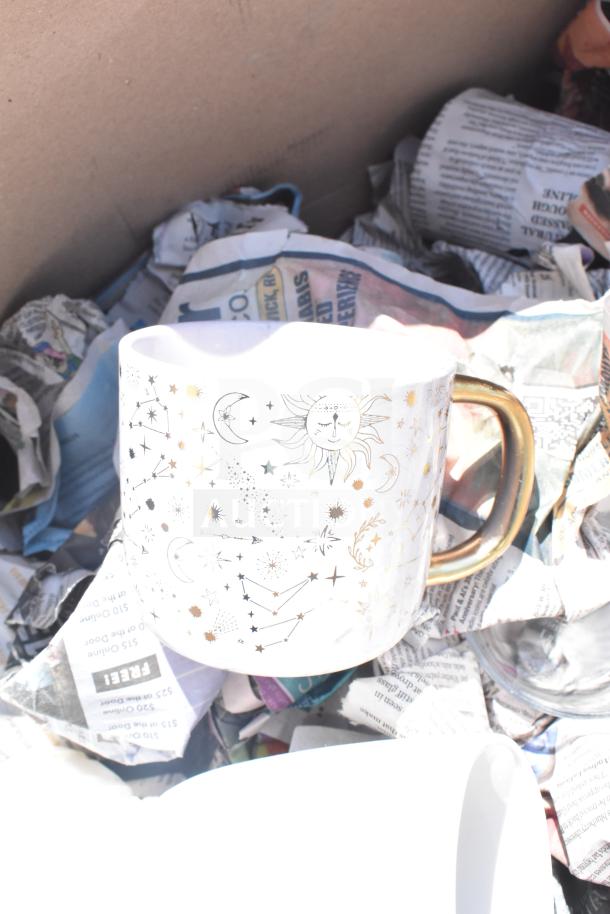 Box containing a celestial-themed mug with gold handle, wrapped in newspaper, part of a lot including glasses and plates.