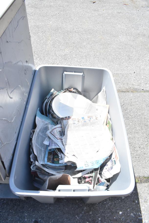 Plastic bin filled with various items including wrapped plates and newspapers. Auction includes two boxes, 2x your bid.