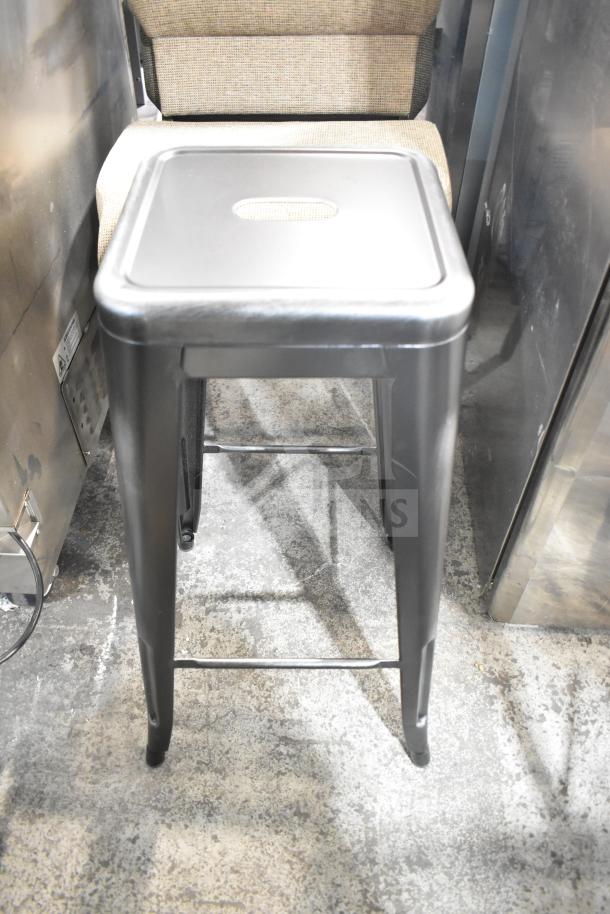 Bar stool with a metal frame and a flat seat, shown in an industrial setting. Includes upholstered chair in background.
