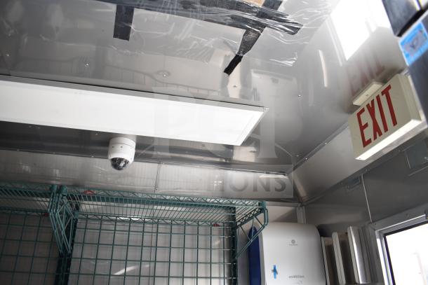 Interior view of a 2021 Texino food trailer, showing ceiling light, security camera, wire shelving, and exit sign.