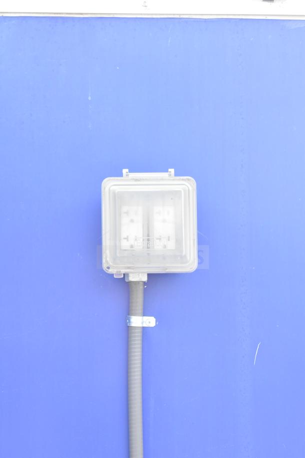 Electrical outlet on a blue surface, encased in a clear box labeled "Extra Duty," connected to a gray conduit.