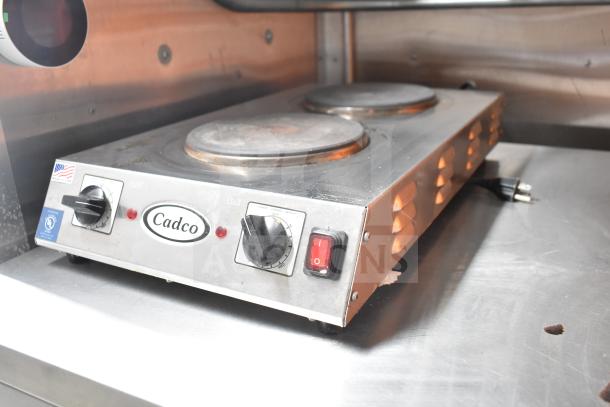 Cadco dual-burner electric hotplate on stainless steel counter in ghost kitchen trailer. Features knobs and power switch.