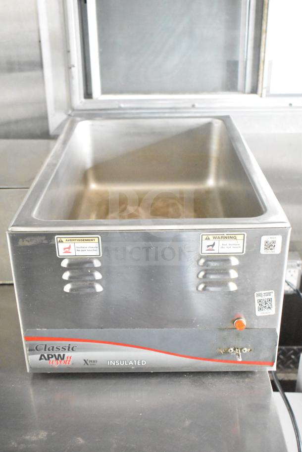 Industrial food warmer with stainless steel construction, visible warnings for hot surface, branded APW Wyott on the label.