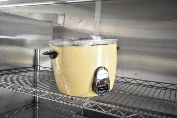 Yellow rice cooker on a metal shelf in a kitchen, covered with plastic wrap. Visible label indicates "RICE MASTER" model.
