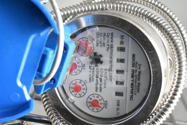 Close-up of an EKM metering water meter, model SPWM-075-CF, with red dials and blue protective cover.