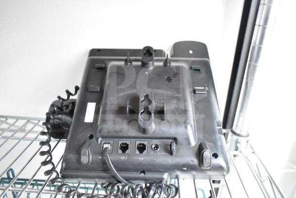 Back view of black electronic device with coiled cord, multiple ports, and mounting slots, resting on wire shelving.