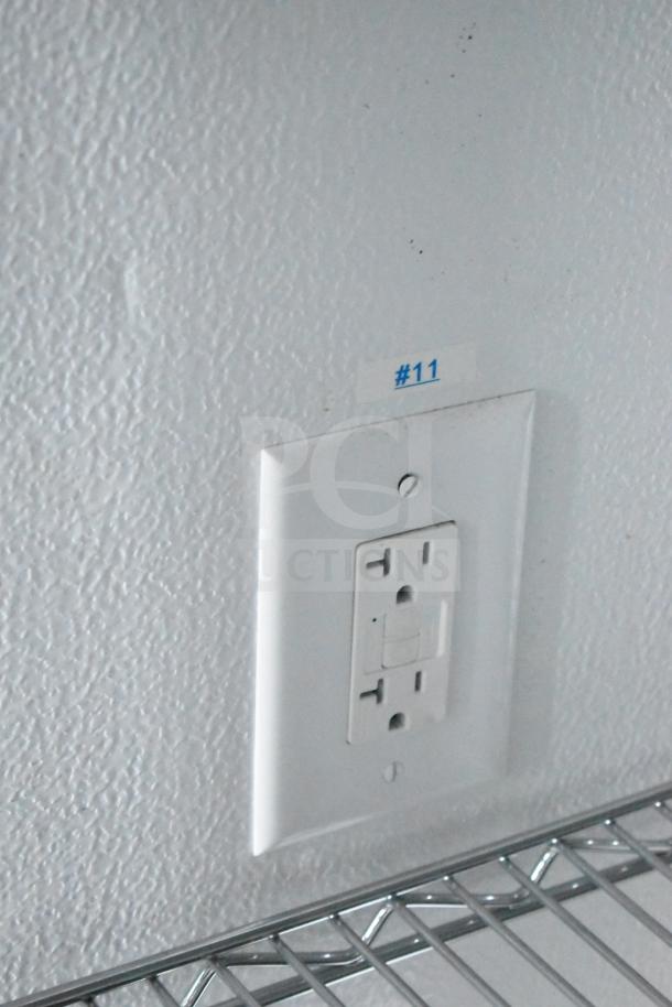 Close-up of an outlet with a "#11" label on a textured wall above a wire shelf inside a mobile commissary trailer.
