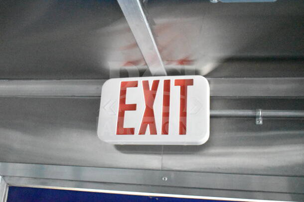 Ceiling-mounted illuminated exit sign in a commercial space, featuring bold red lettering and directional arrows.