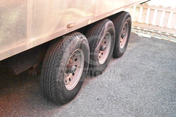 Triple-axle wheels of a 2021 Texino 28ft cooking trailer on asphalt, showing sturdy tires and metal rims.