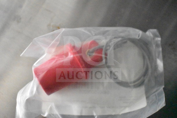 Red safety cap in plastic packaging with a cable loop, shown on a flat surface.