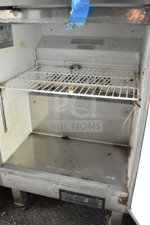 Open interior of a used food trailer refrigerator with wire racks and signs of wear. Manufacturer label visible on the side.