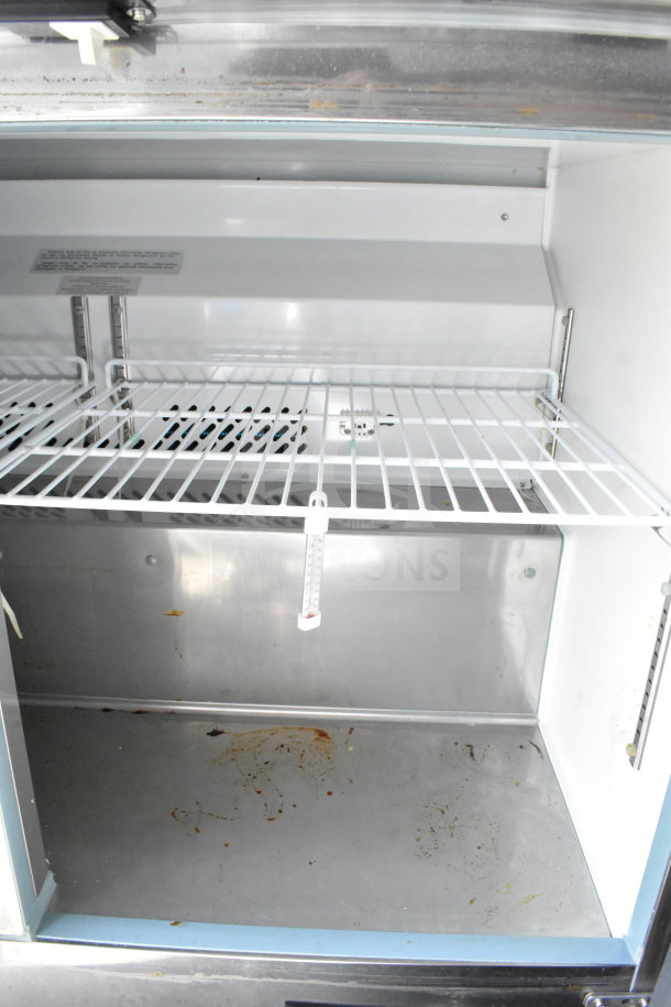 Interior view of a food trailer's refrigerator, showing wire shelving and a thermometer. Some stains are visible on the metal surface.