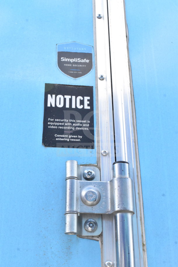 Close-up of a blue Diamond Cargo trailer door showing a security notice about audio and video recording devices, with SimpliSafe security label and metal hinge.