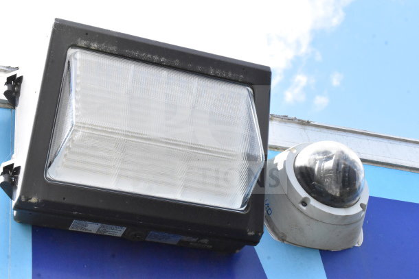 Outdoor security camera and rectangular light fixture mounted side by side on a blue background.