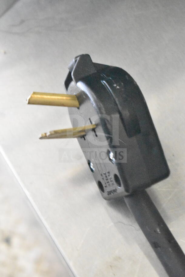 Close-up of a plug from a 2021 Diamond Cargo 8.5'x24' food trailer. Black with brass prongs, resting on metal surface.