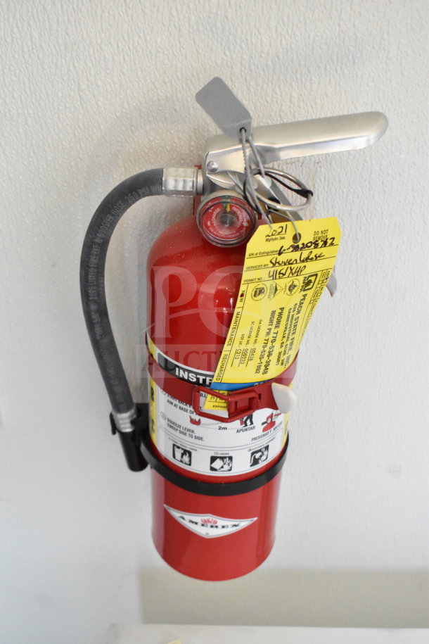Red fire extinguisher with inspection tags attached and pressure gauge visible, mounted on a white wall.