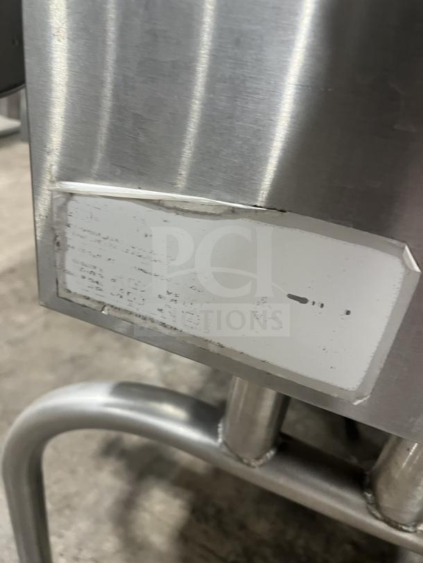 Commercial stainless steel braising pan on sturdy legs, slight wear. Features tilting mechanism. Indistinct label visible.