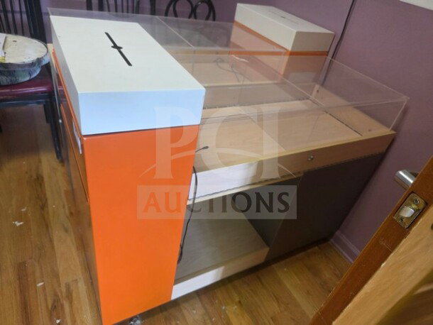 Modern retail display checkout counter with orange and white panels, clear acrylic top, and drawer. Slight wear visible.