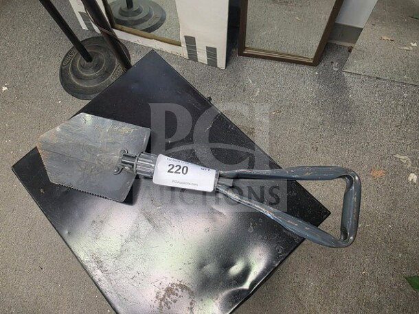 Vintage entrenching tool with a collapsible metal handle, serrated edge, and label marked "220 PCUauctions.com." Signs of wear visible.