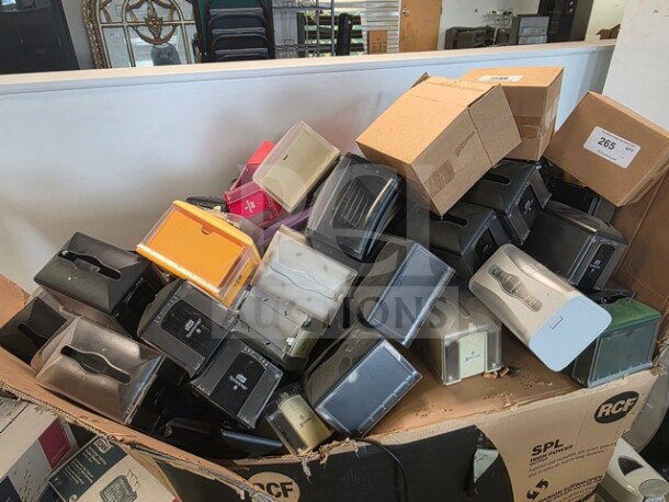 A large collection of assorted napkin dispensers piled in an open cardboard box, various colors and styles visible.