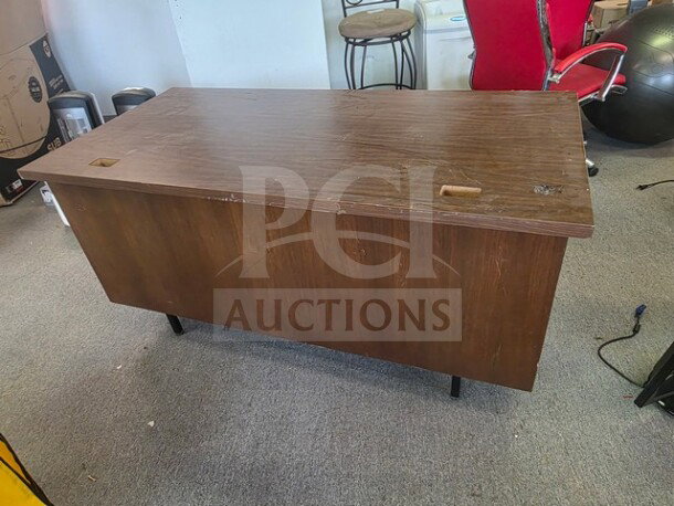 A wooden pedestal desk missing drawers, showing surface wear and scratches, on a carpeted floor in an office setting.