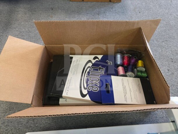 Open box containing an embroidery machine and colorful spools of thread, items enclosed in packaging with visible labels.