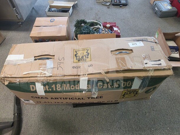 Cardboard box labeled "CMAS Artificial Tree" with assorted Christmas decorations inside, including greenery and red boxes. Box shows signs of wear, taped handles visible.