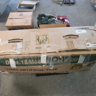 Cardboard box labeled "CMAS Artificial Tree" with assorted Christmas decorations inside, including greenery and red boxes. Box shows signs of wear, taped handles visible.
