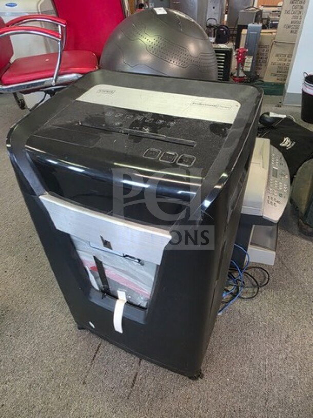 Black paper shredder with visible control panel and paper slot. Behind it, a fax machine and printer are partly visible.