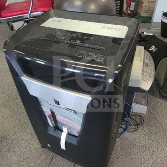 Black paper shredder with visible control panel and paper slot. Behind it, a fax machine and printer are partly visible.