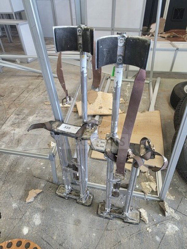 A pair of well-used drywall stilts with adjustable straps, metal construction, and padded leg supports, item tag 335 visible.