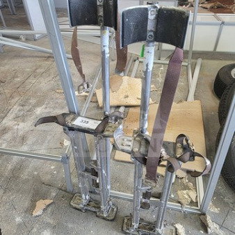 A pair of well-used drywall stilts with adjustable straps, metal construction, and padded leg supports, item tag 335 visible.