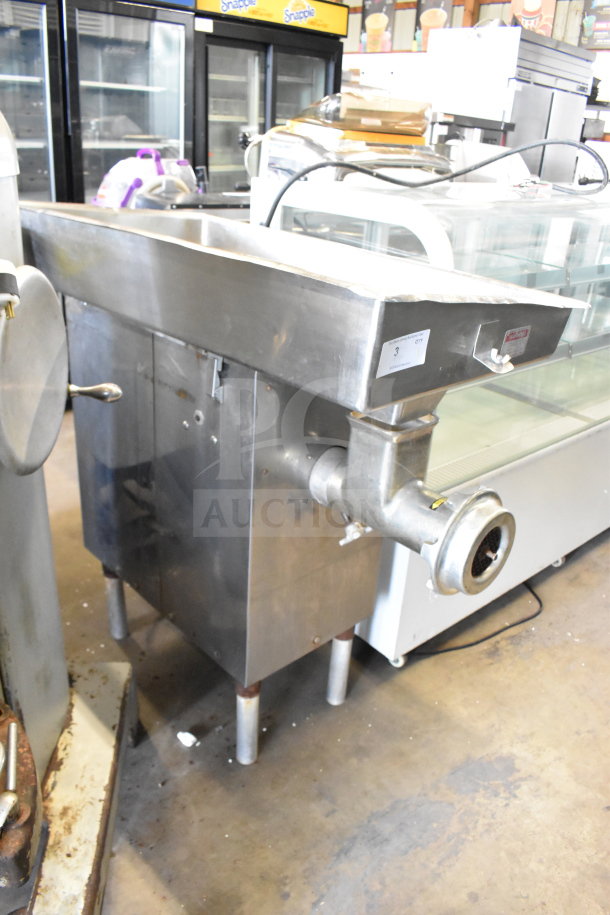 Commercial floor-style meat grinder in stainless steel, shows wear. Includes collection tray and grinding head.