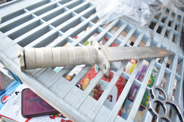 A serrated knife with a textured handle is displayed on a metal grate, surrounded by colorful packaging and black knuckles.