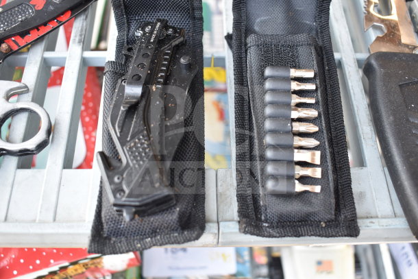13 various items including a black folding knife, chrome knuckles, and a set of drill bits. Black fabric cases visible.