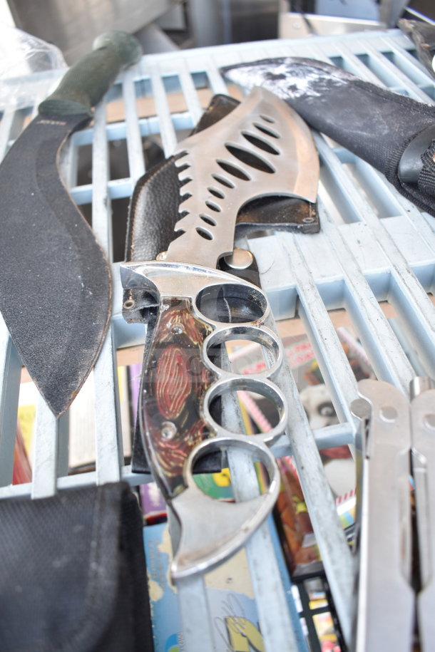 Assorted items including knives, knuckles, and drill bits on a table. Some knives have unique blade designs. Items are in used condition.