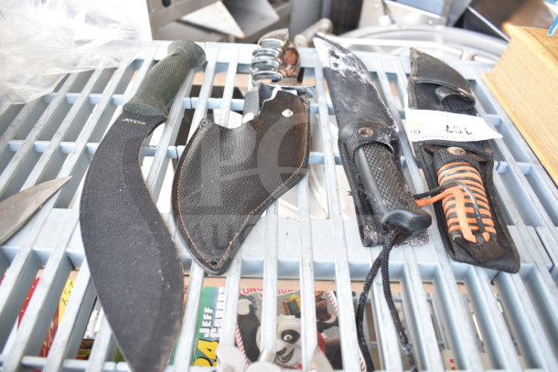 Various knives and knuckles on display, including a machete and tactical knife with sheaths, on a metal grid. Auction tag visible.