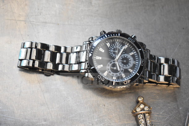 Silver-tone chronograph wristwatch with black dial and gem markers, alongside a small, jeweled brooch.