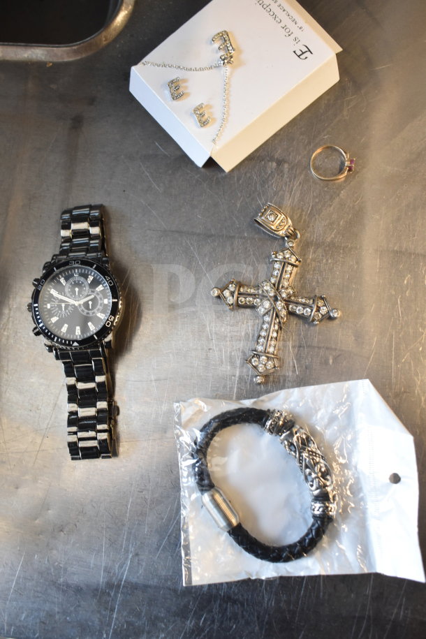 Case of assorted jewelry including a black watch, ornate cross pendant, ring, "E" earrings on card, and a braided bracelet.