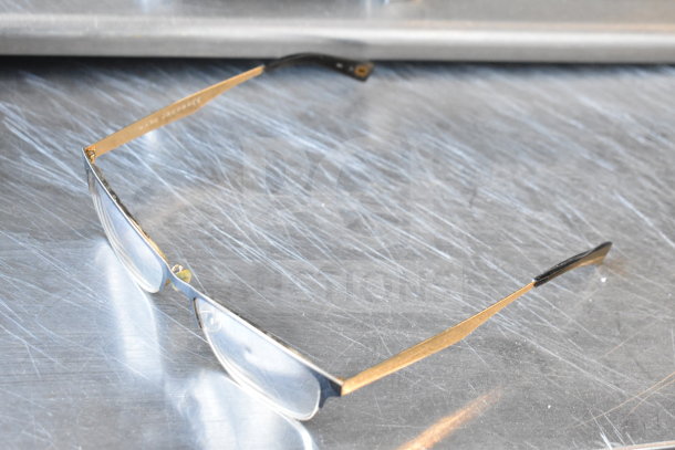 Rectangular eyeglasses with gold arms and black tips, displayed on a metallic surface. Left arm inscribed "Marc Jacobs".