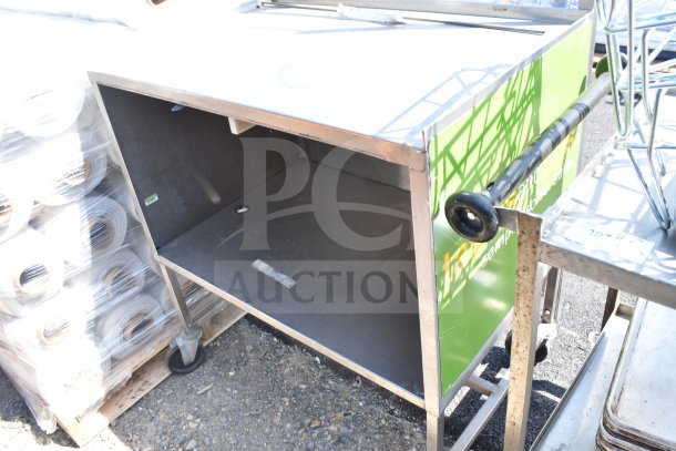 Commercial table with under shelf, stainless steel, on commercial casters, showing some wear, partially visible green panel.