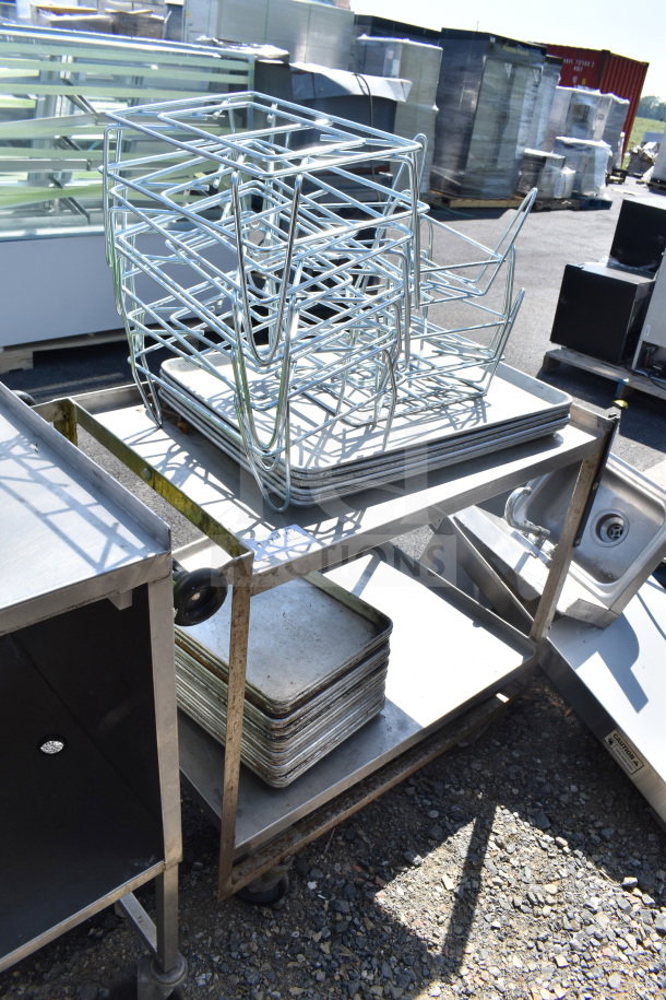 2 tier metal cart with wheels, holding metal baking pans and silver racks, in used condition outdoors.
