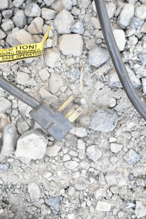 Electrical plug on rocky ground, connected to a cable. Yellow caution tag reads: "Ground required for safe operation."