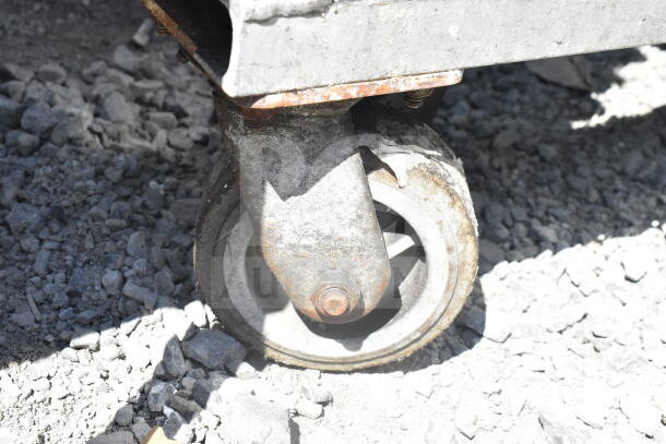 Rusty caster wheel attached to a commercial pan transport rack on gravel surface.