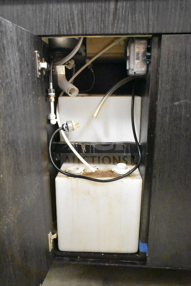 Portable bar storage compartment showing plumbing fixtures and a white storage tank inside a dark cabinet.