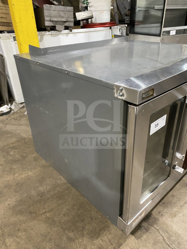 Industrial stainless steel refrigeration unit with glass front, visible wear, and manufacturer label.