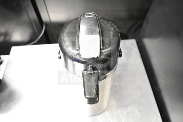 Stainless steel air pot with black top, showing signs of use. Features a hinged lid and dispensing spout.